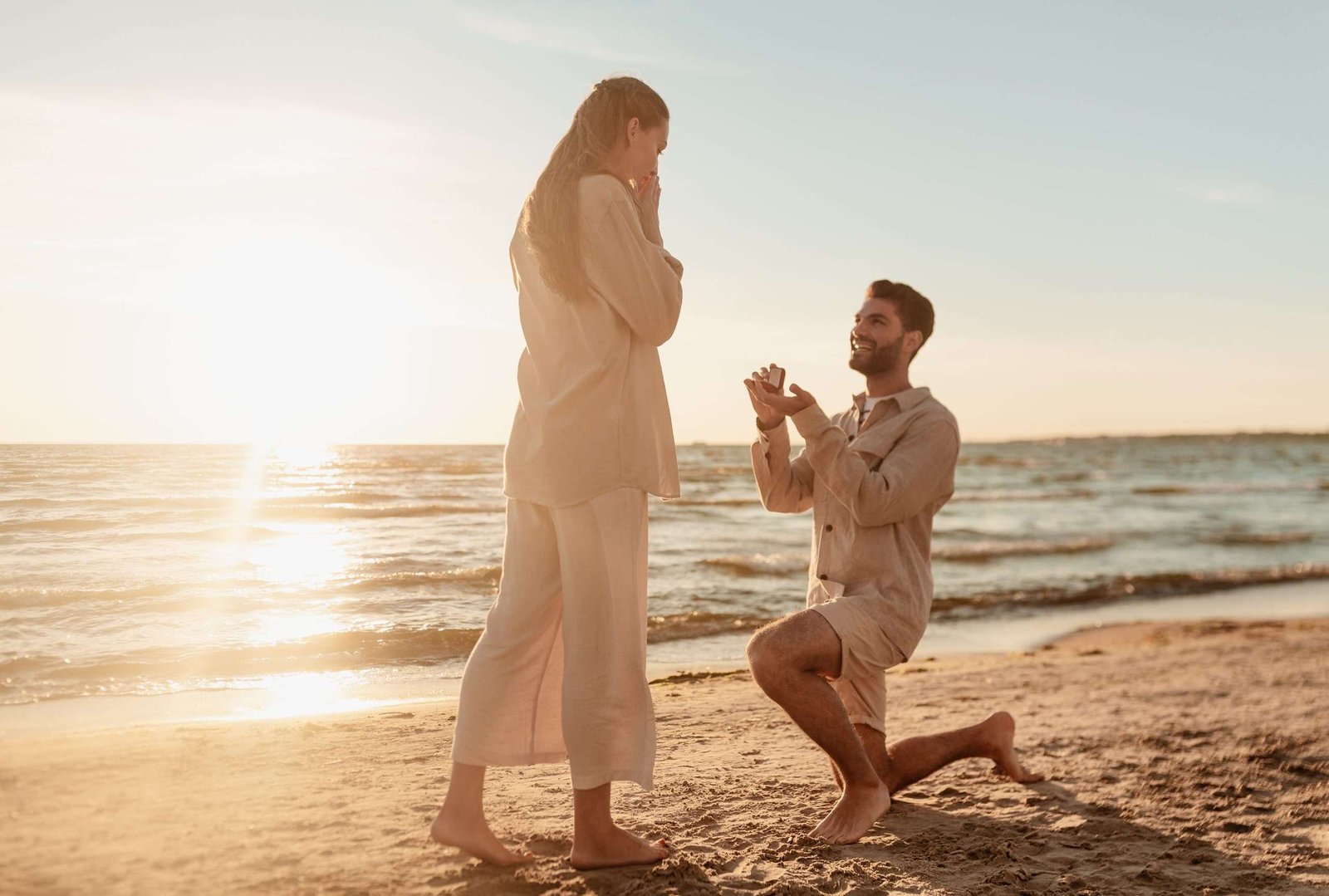 smiling young man with diamond engagement ring making proposal to happy woman on beach